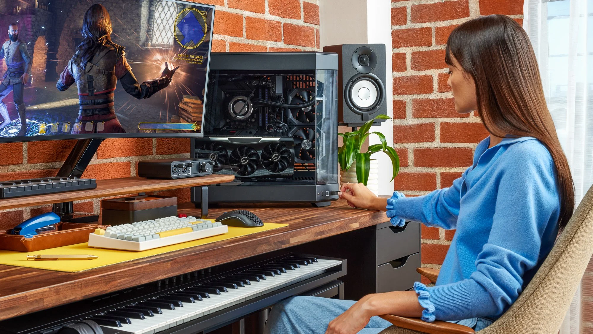 A woman sitting at a desk in front of a computer setup and a large monitor playing a video game, with a keyboard, mouse, and sound equipment on the desk, and a brick wall in the background.