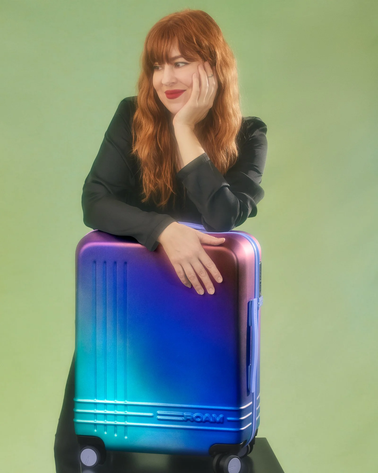 A woman with red hair, wearing a black satin jacket, resting her head on her hand, leaning on a colorful, gradient rolling suitcase against a green background.