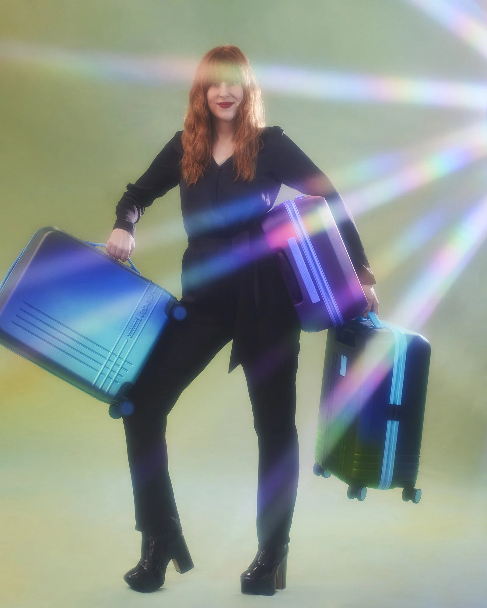 A woman with long red hair, wearing a black outfit and high heels, holding three colorful suitcases against a pastel background with rainbow light effects.