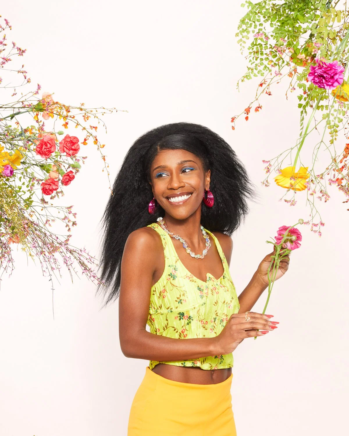 A woman with dark, frizzy hair smiling and holding a pink flower, surrounded by colorful flowers against a white background.