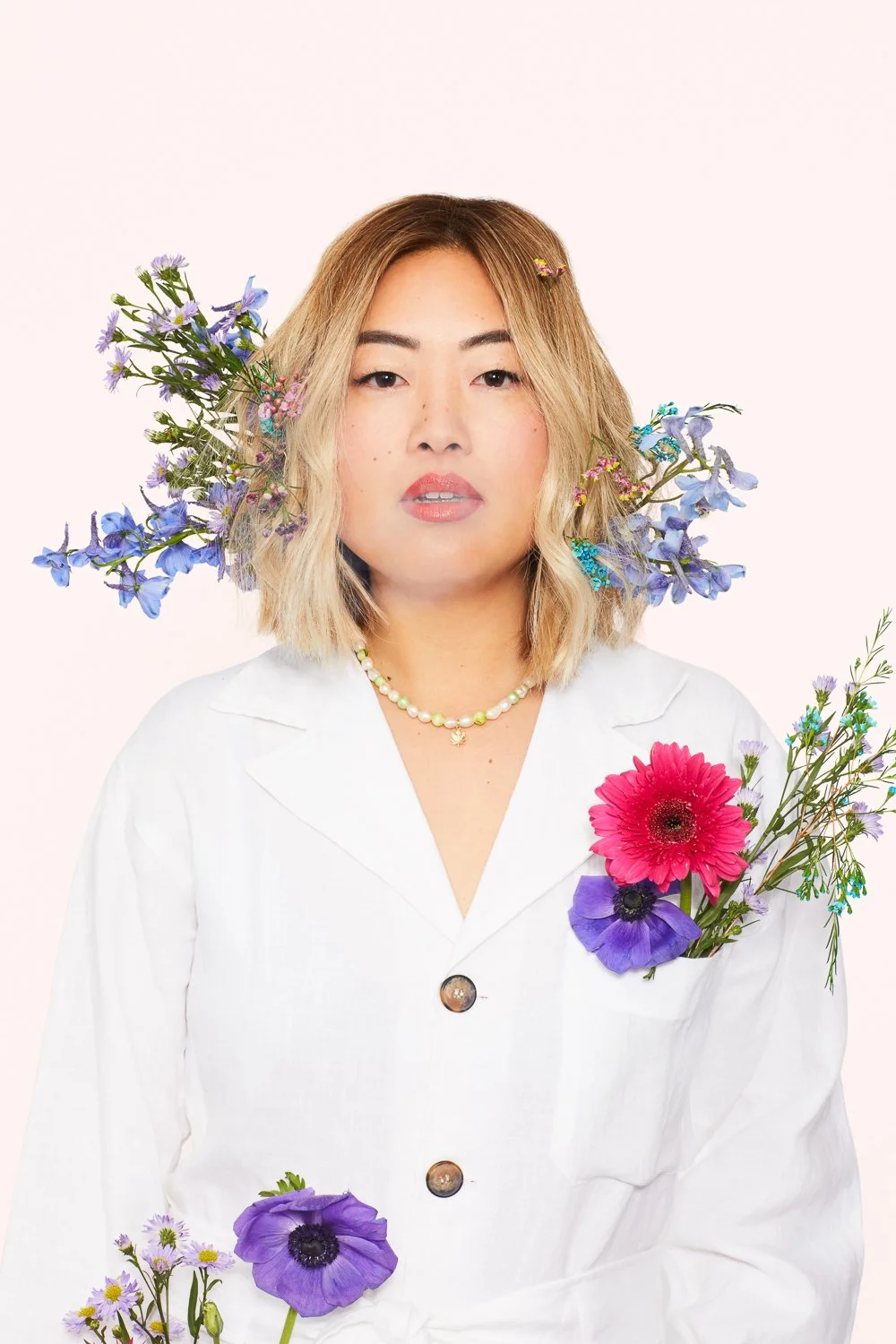 A woman with shoulder-length blonde hair wearing a white blazer and pearl necklace, surrounded by colorful flowers including purple, pink, and blue blossoms.