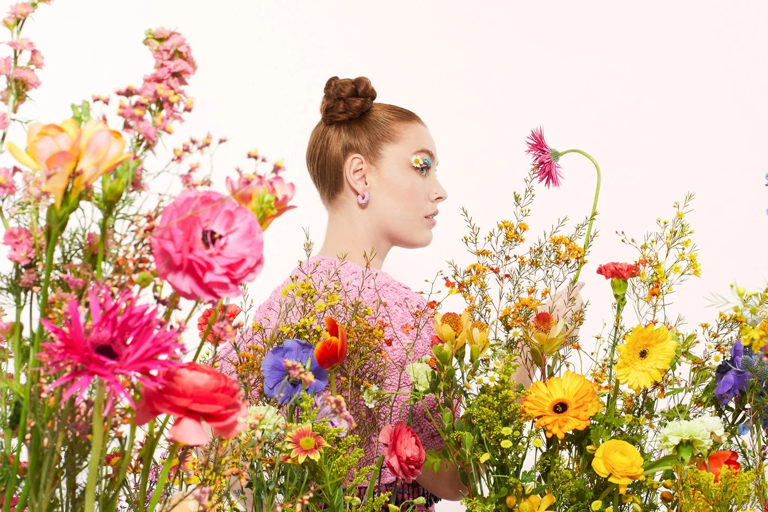 A woman with styled hair and floral makeup surrounded by colorful flowers, holding a pink flower, against a plain white background.