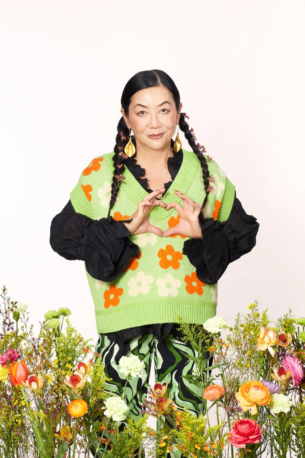A woman with black hair styled in braids, wearing large gold earrings, a black blouse with ruffled collar, a green sweater vest with orange and white flowers, and black and green patterned pants, standing behind colorful flowers making a heart shape 