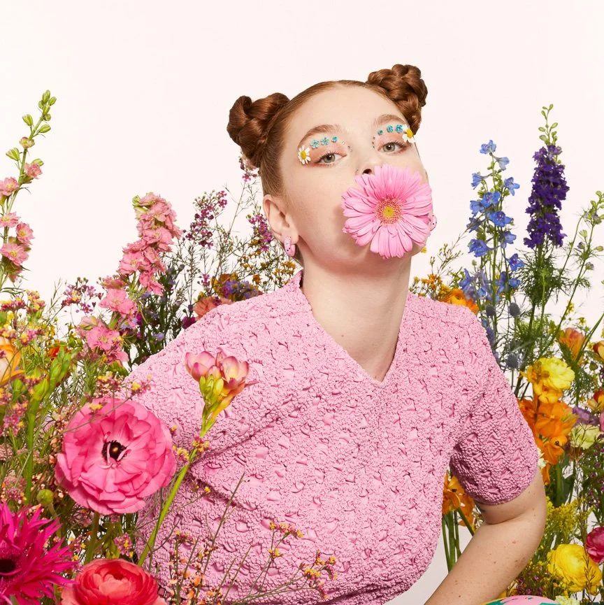 A woman with red hair styled in two buns, wearing pink clothing, surrounded by colorful flowers, with a pink flower covering her mouth and decorated with small flowers and daisies around her eyes.