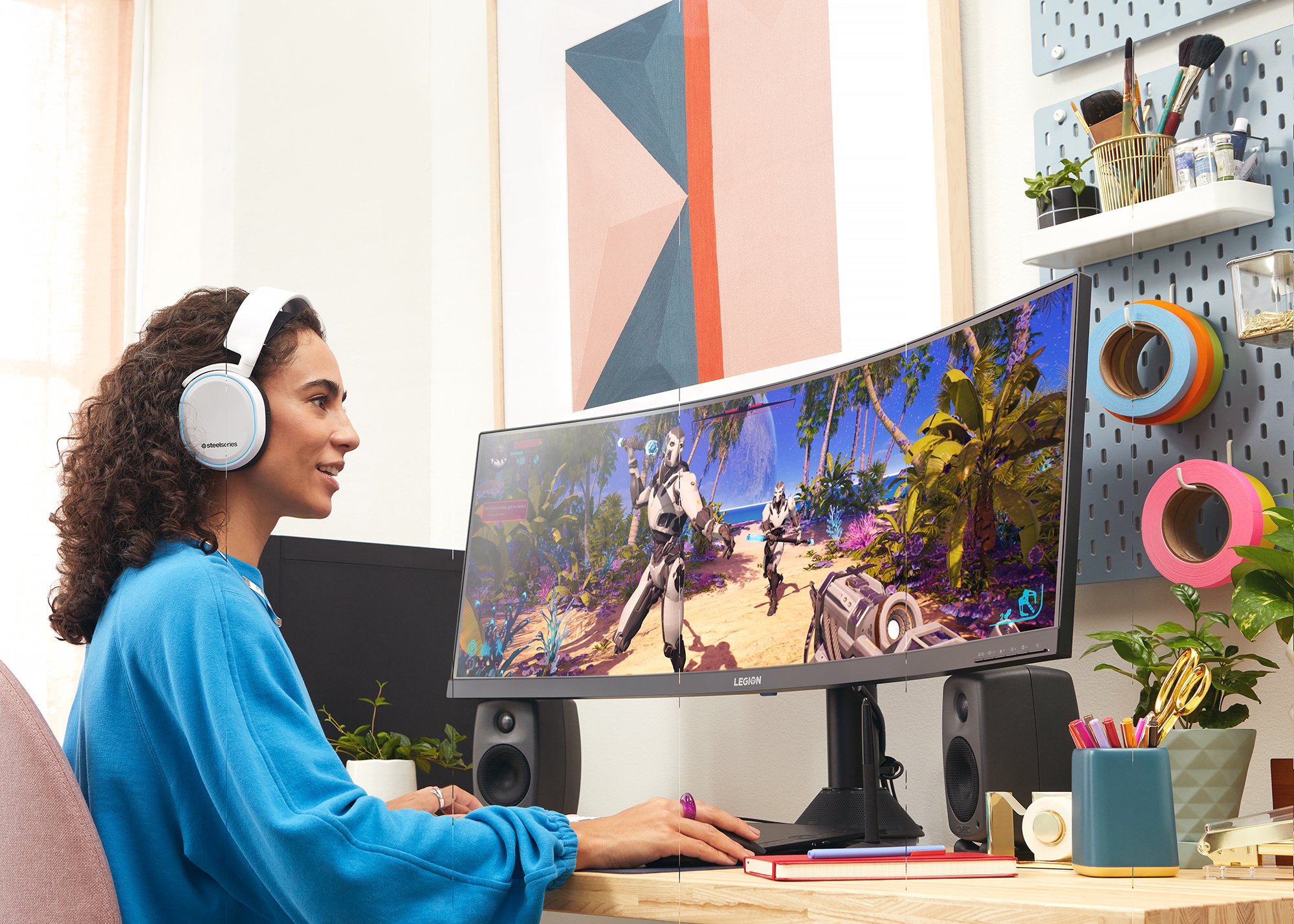 A woman with curly hair wearing a blue sweatshirt and white headphones, sitting at a desk with a curved monitor displaying a video game with robots on a tropical island. The desk has plants, writing utensils, and a pegboard with art supplies and deco