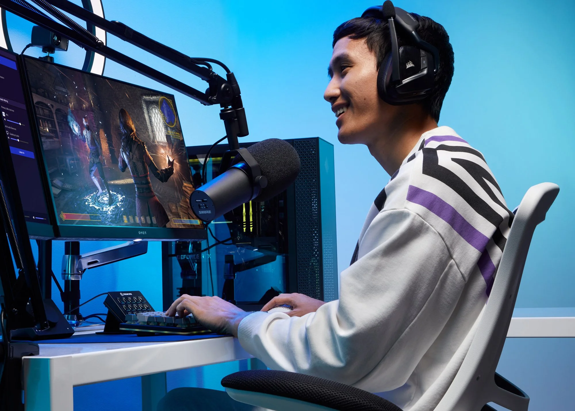 A young man with headphones is playing a video game on his computer, captured in a side profile shot. He is smiling and sitting at a white desk with a monitor displaying a fantasy game scene.