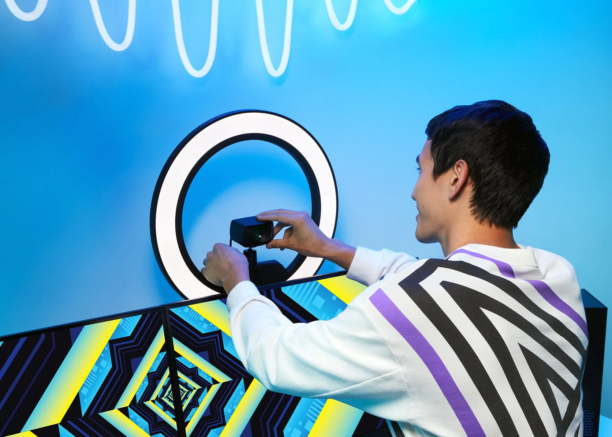 A man with dark hair taking a photo of a geometric, optical illusion art piece with a camera, in a brightly colored room with blue walls and neon light decorations.