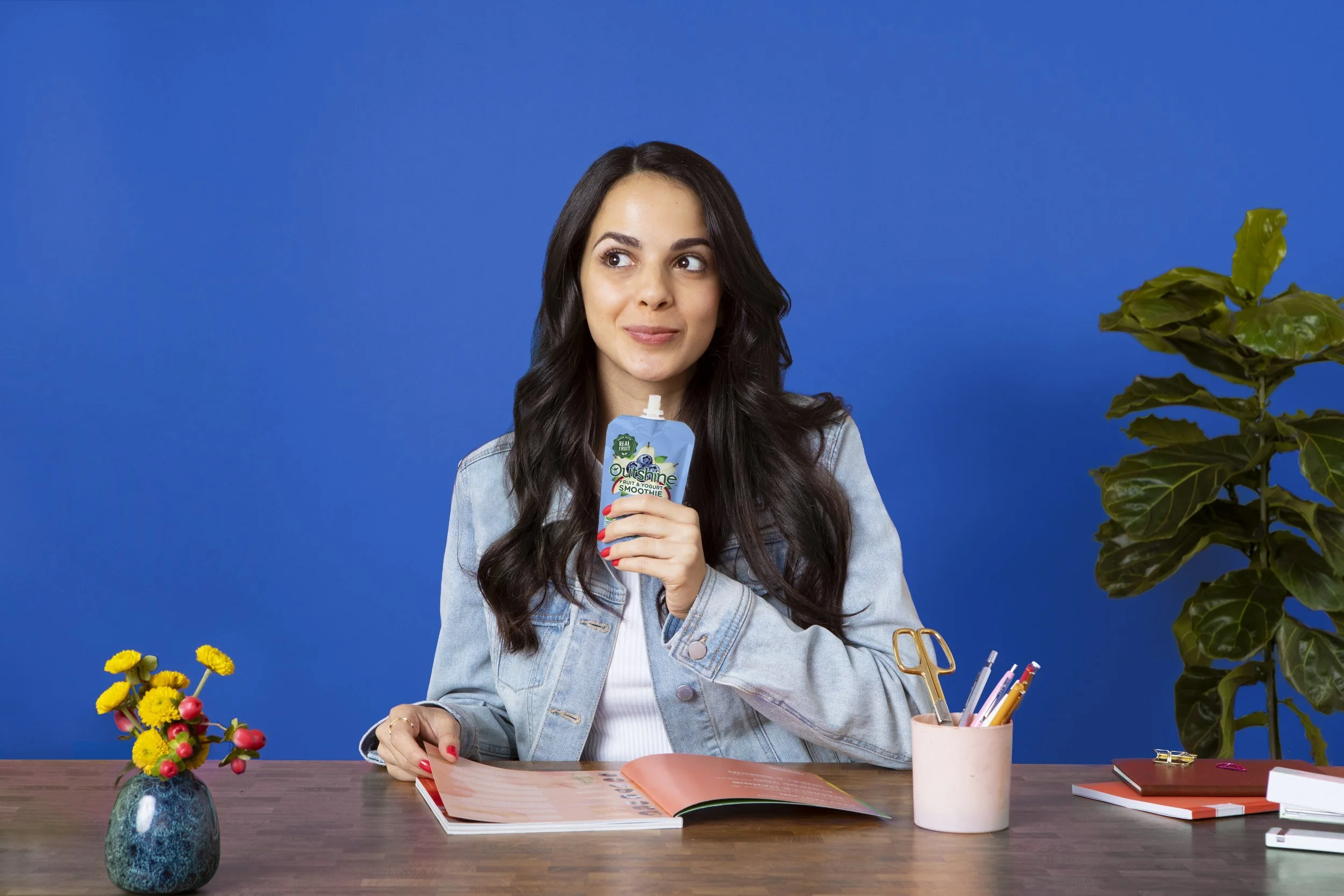 Young woman with long dark hair sitting at a desk with a blue background, holding a squeeze pouch of smoothie, with an open notebook, a pink mug with pens, and a potted plant and flowers on the desk.