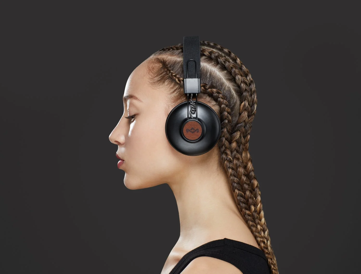 Profile of a young woman with braided hair wearing large black over-ear headphones against a dark background.