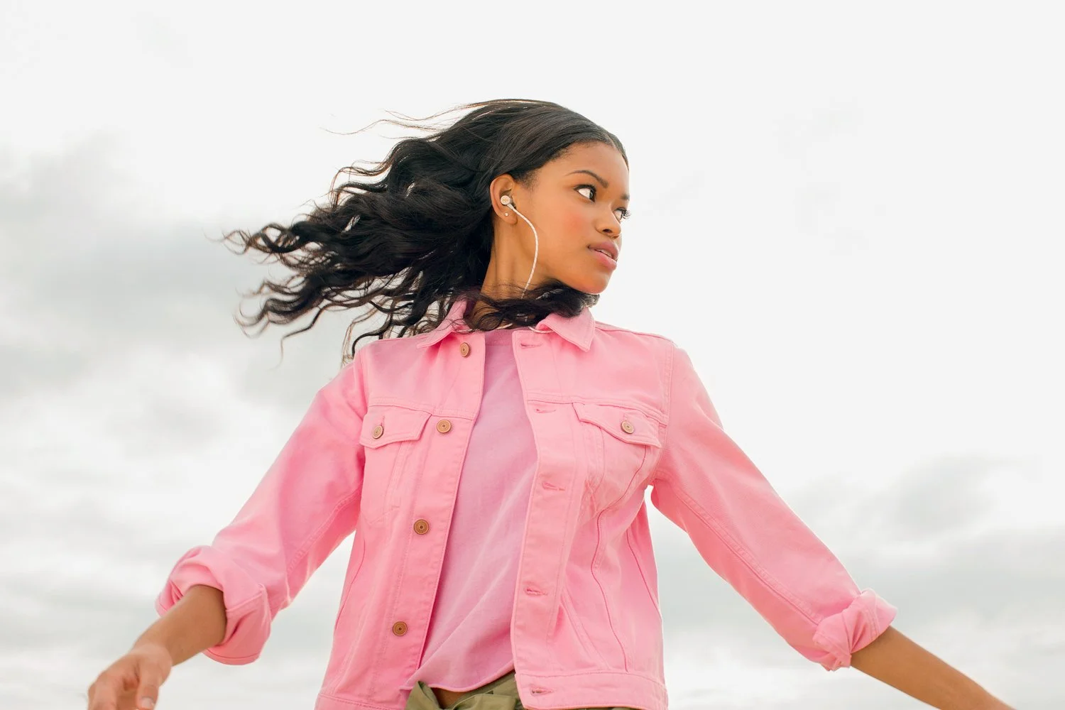 Young woman with long, wavy hair, wearing a pink jacket and earphones, looking to her right outdoors on a cloudy day.