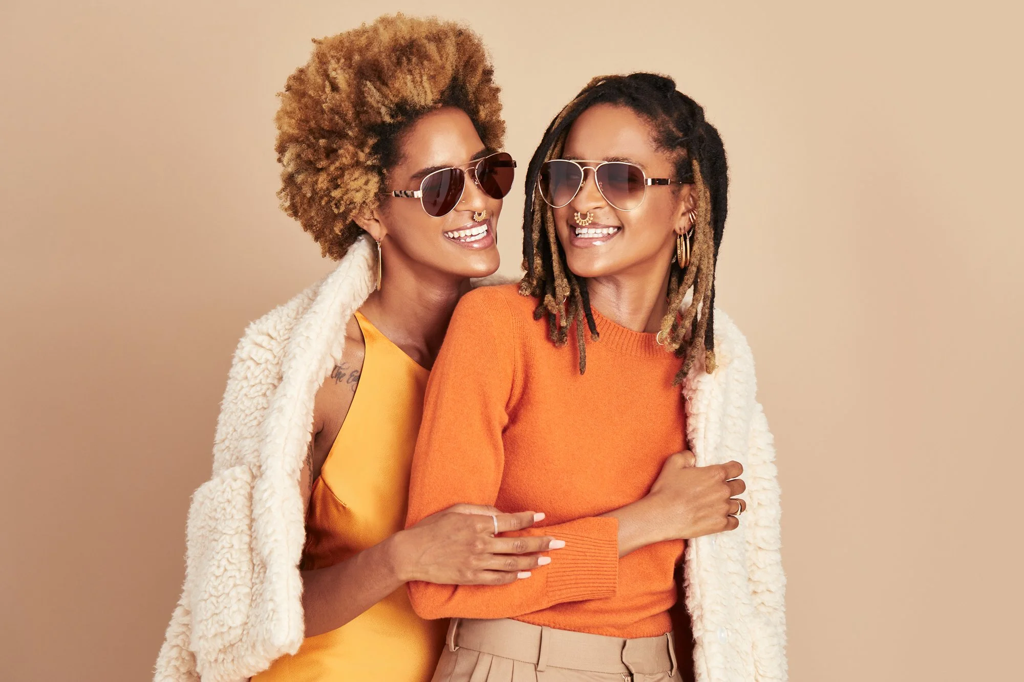Two women with dreadlocks and curly hair smiling and wearing sunglasses, one in an orange sweater and the other in a yellow top, posing together against a beige background.