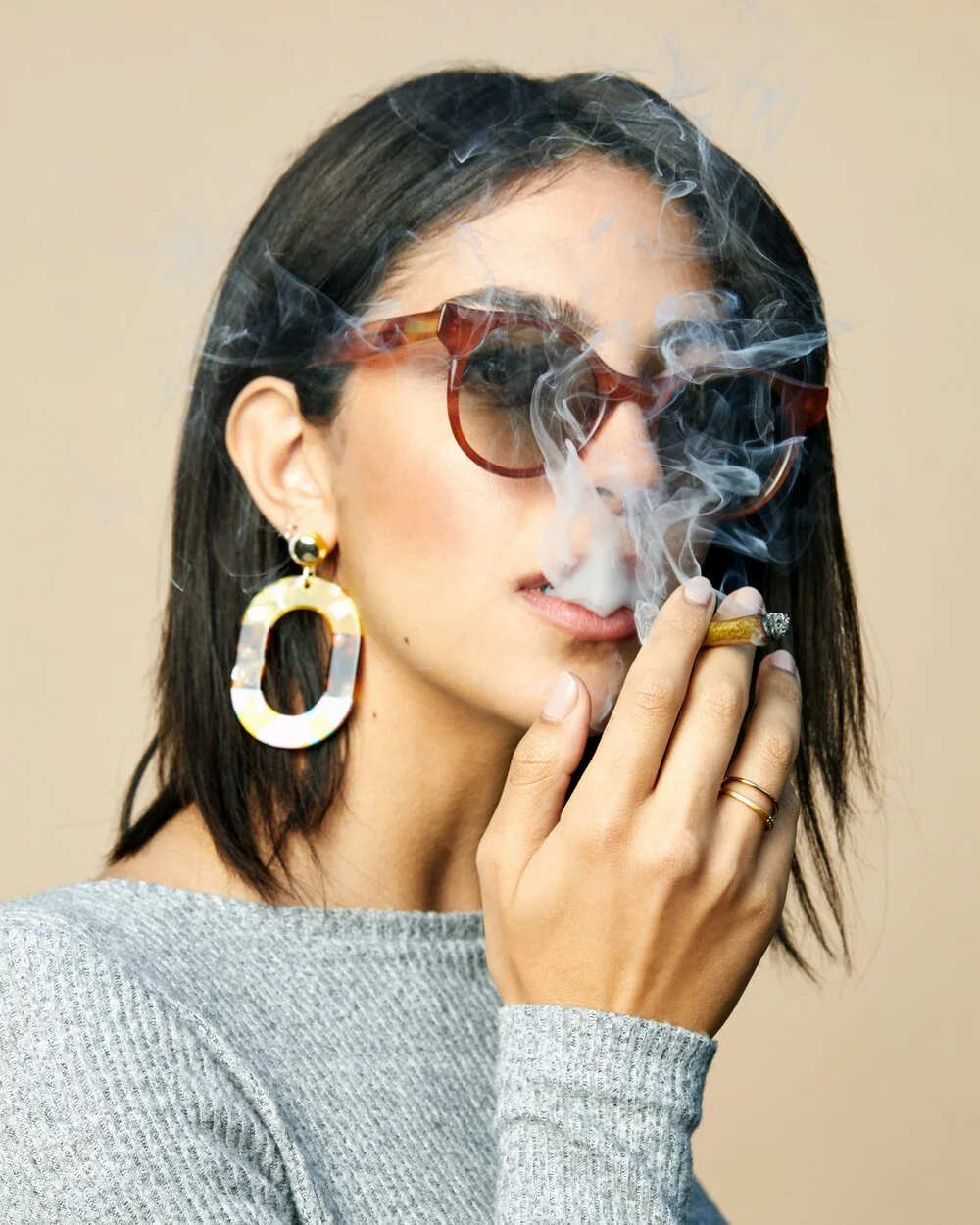 A woman with dark hair, wearing red sunglasses and large gold earrings, is smoking a cigarette with smoke rising around her face.