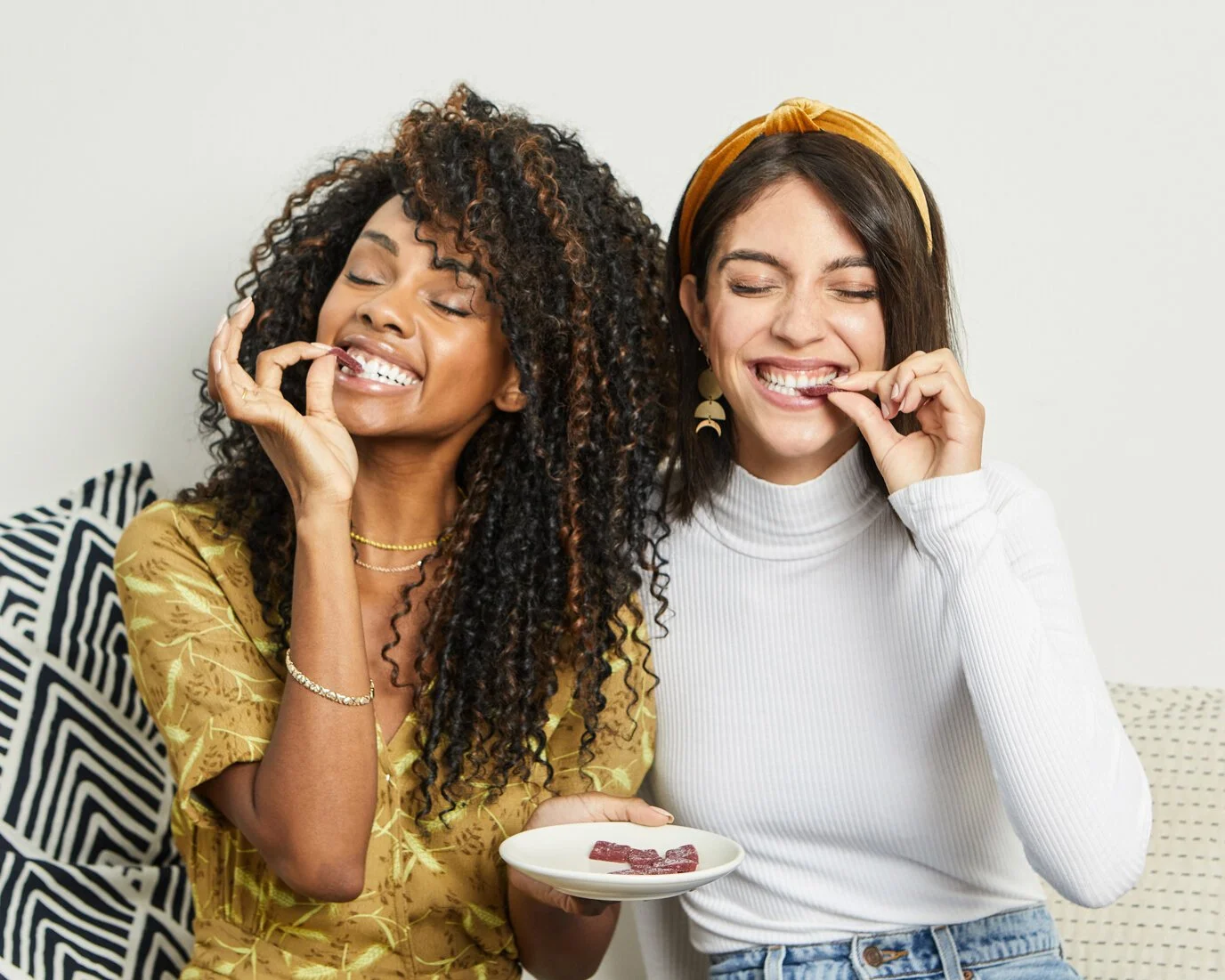 Two women sitting together, enjoying and eating candy from a white bowl, smiling and having a good time.