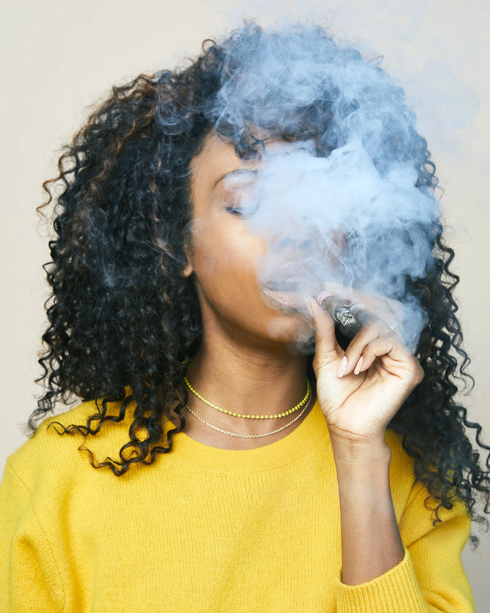 A woman with curly hair wearing a yellow sweater and layered necklaces, smoking a cigar with smoke covering her face.