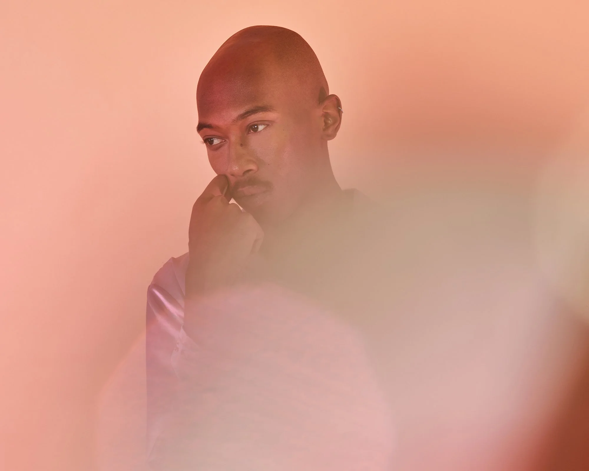 A young person with a shaved head looking to the side with hand near face, set against a muted pink background.