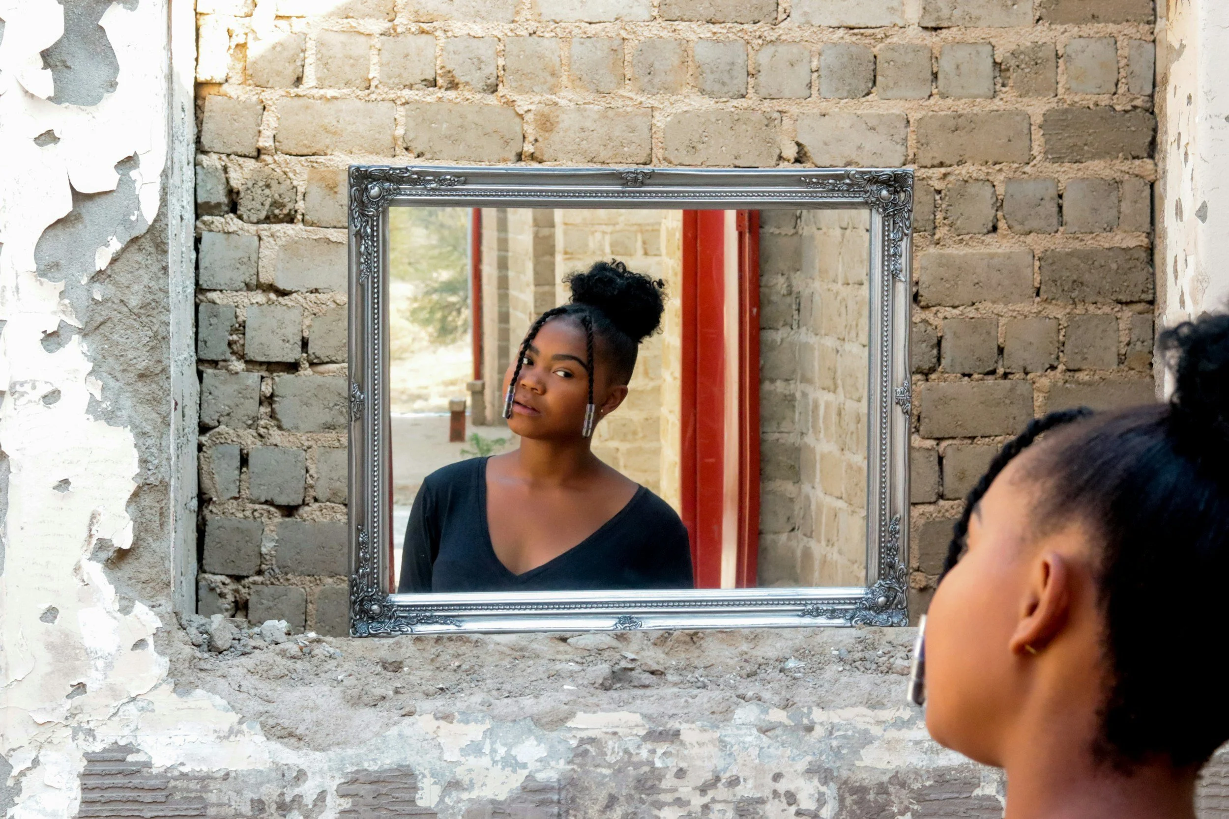 A black woman with braided hair and earrings looking into a mirror set in a brick wall, with her reflection visible in the mirror.