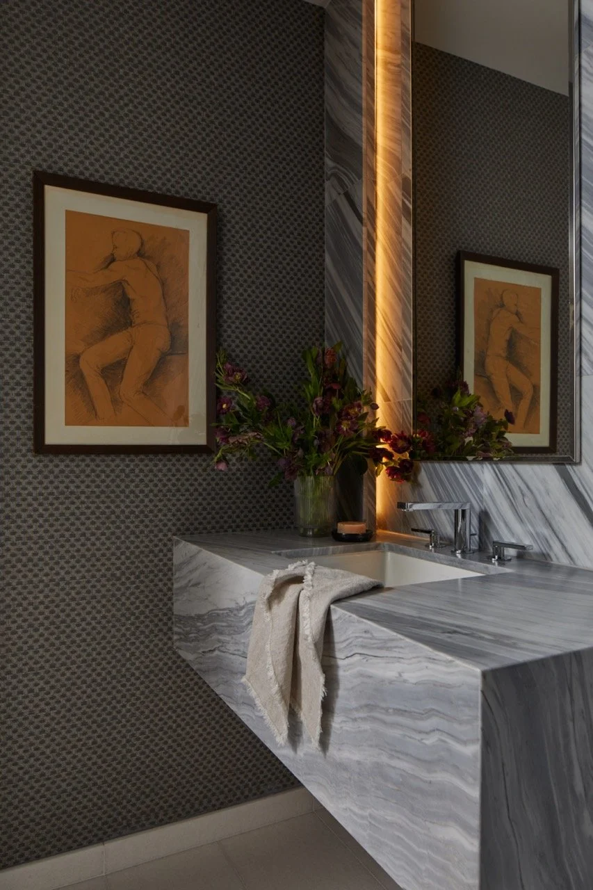 Modern bathroom with marble countertop, underlit mirror, dark gray textured wallpaper, framed sketches, vase with flowers, and towel on the sink.