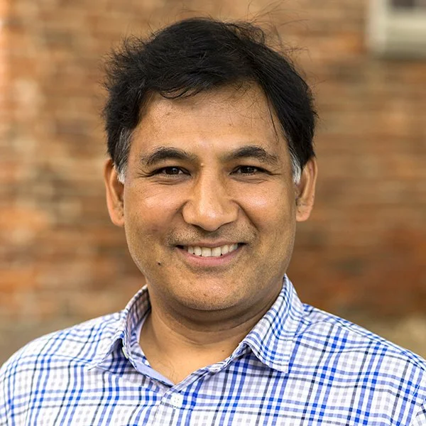 A man with dark hair, smiling, wearing a blue and white checkered shirt, standing outdoors with a brick wall in the background.