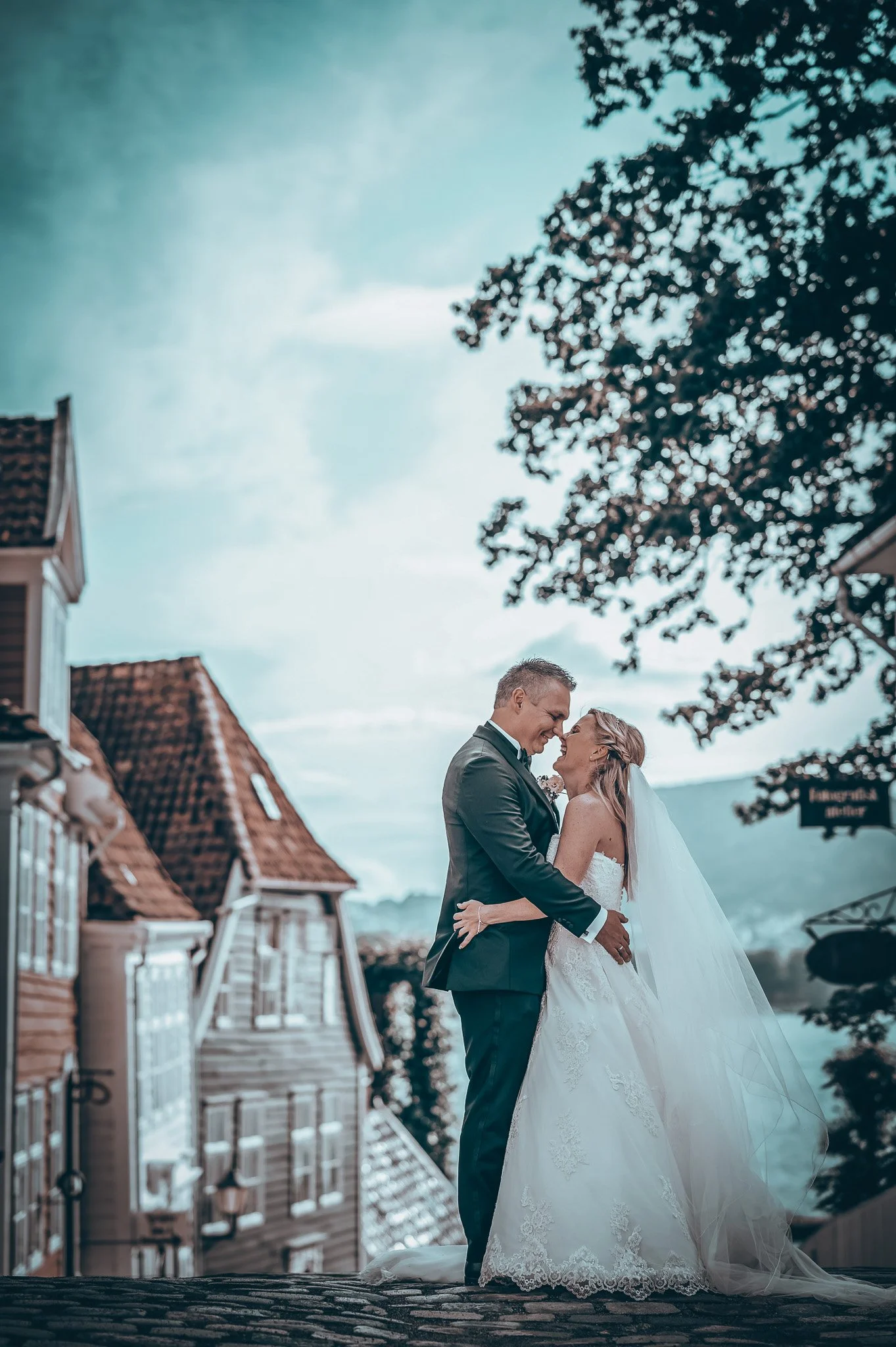 Bryllupsfoto/video: Brudepar som omfavner hverandre foran gamle bygninger med teglstein i bakgrunnen. Bruden har på seg en hvit kjole og brudgom en mørk dress. Det er en klar himmel og noen trær i nærheten.