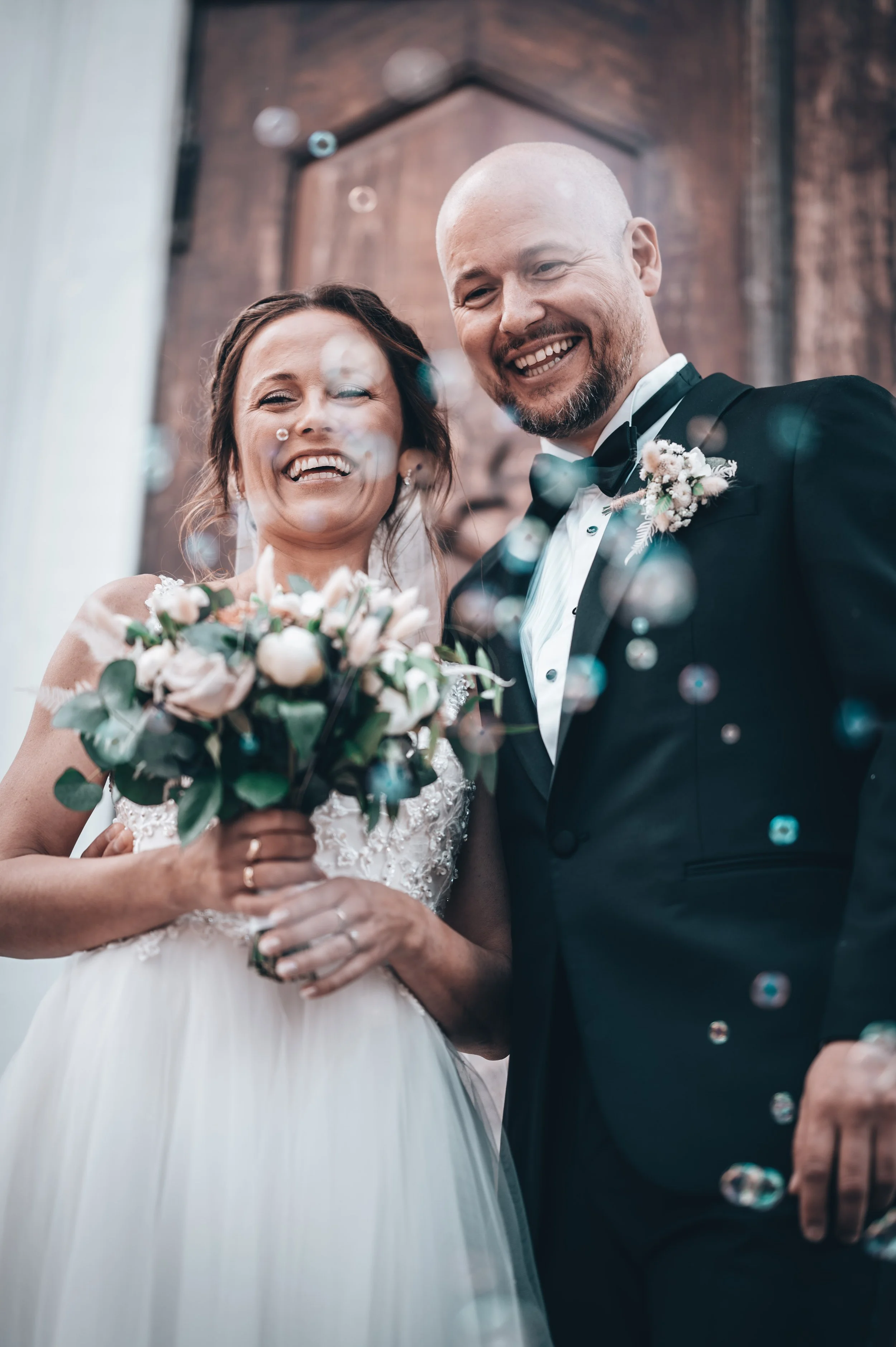 Bryllupsfoto/video: Brud og brudgom smilende, holder en blomsterbukett, omgitt av såpebobler.