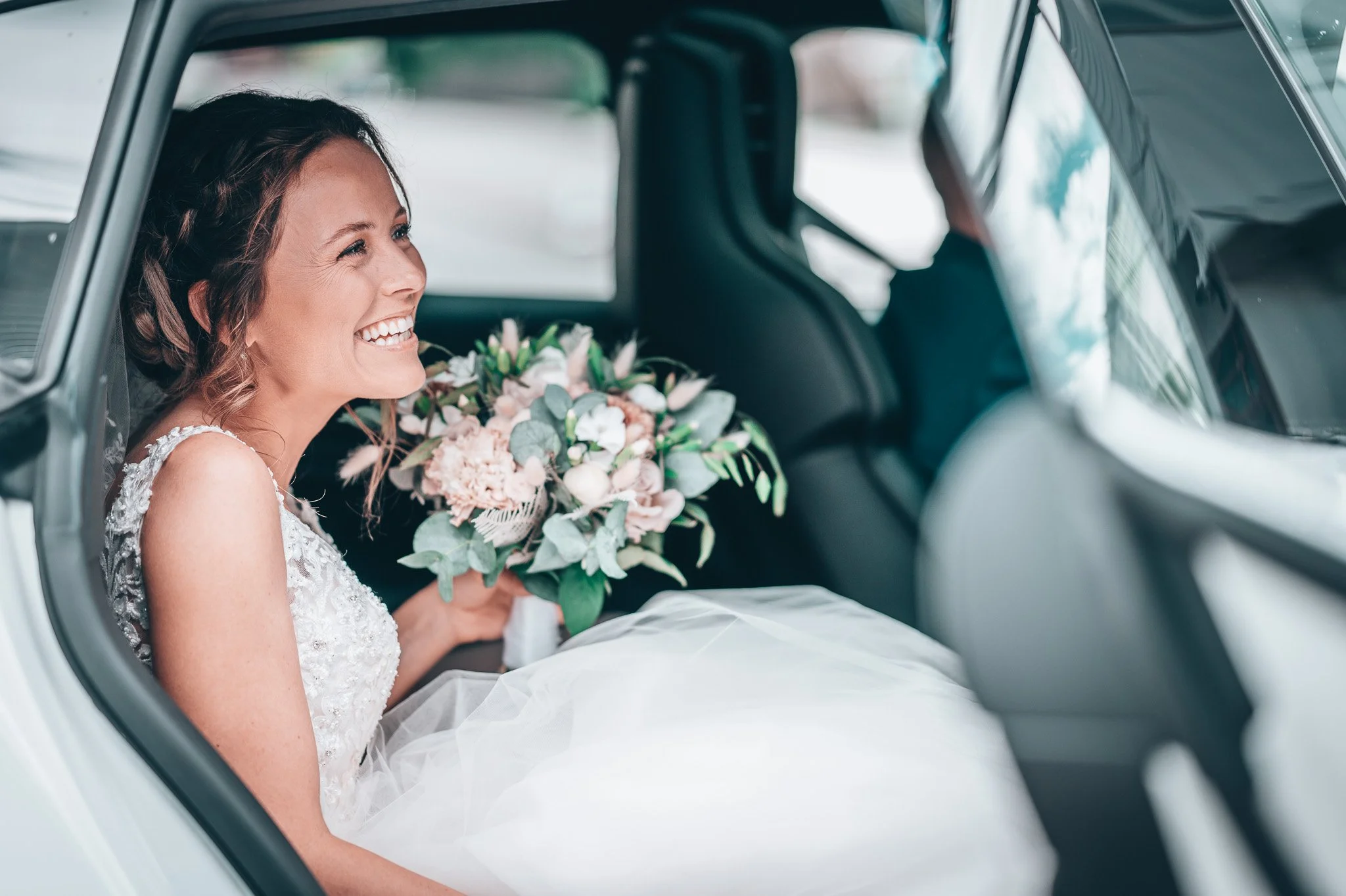 Bryllupsfoto/video: Brud som sitter i en bil og holder en blomsterbukett, smilende i en hvit brudekjole.