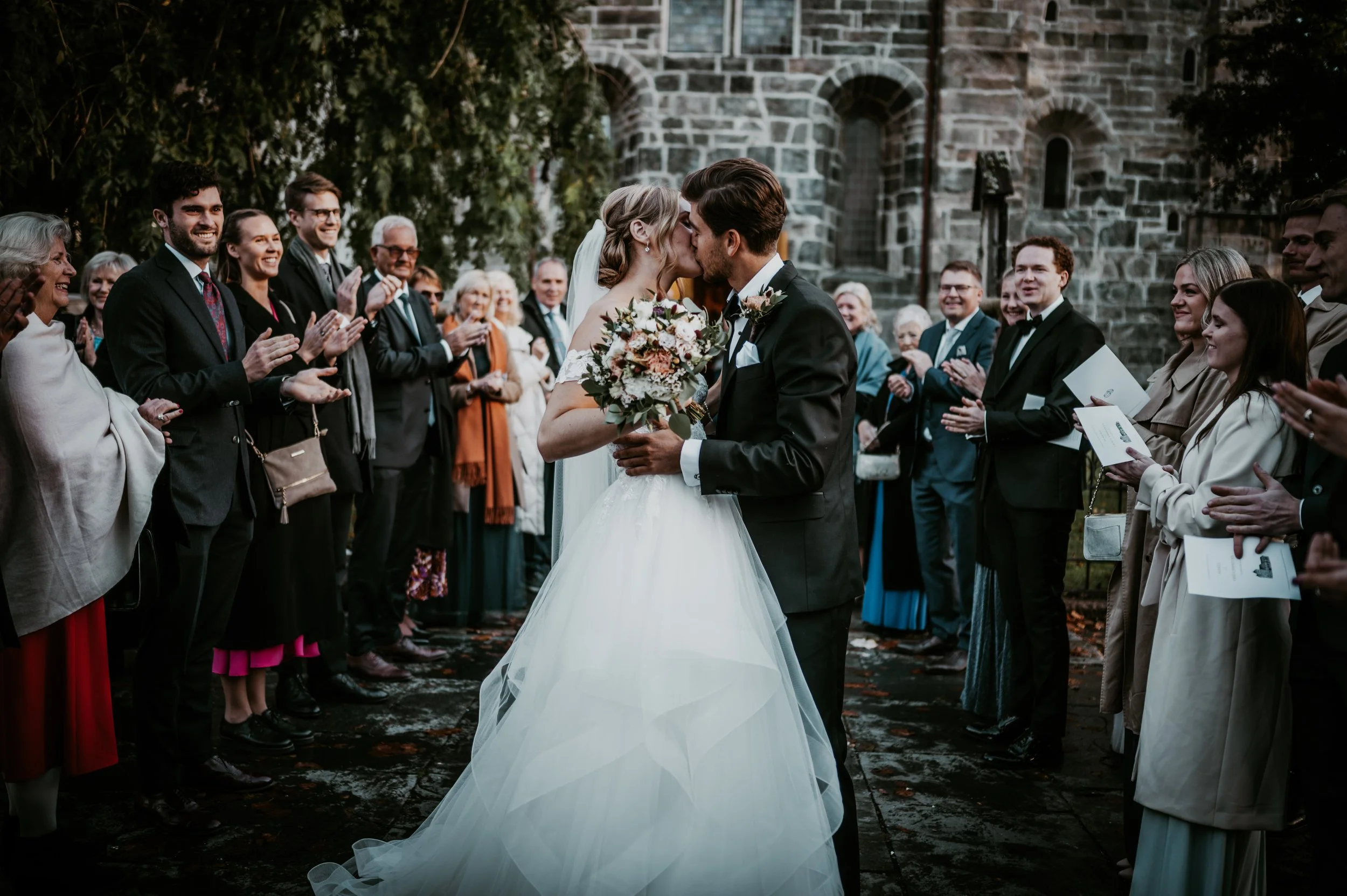 Bryllupsfoto/video: Brudepar kysser foran en gruppe klappende gjester utenfor en steinkirke. Brudens kjole er hvit og brudgommen har på seg en mørk dress. Gjestene er kledd formelt og smiler.