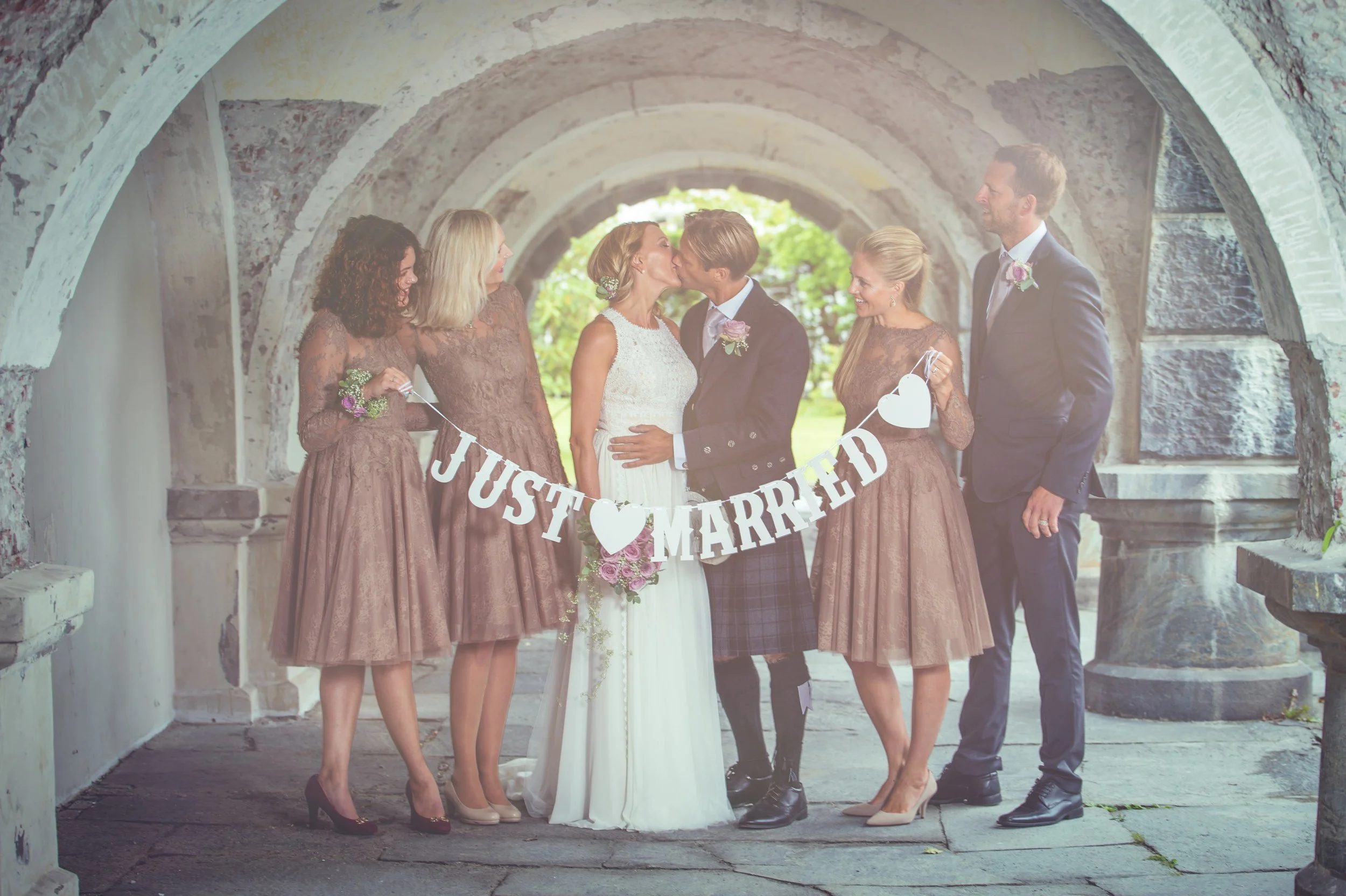 Bryllupspar som kysser, omgitt av brudepiker og forlover som holder "Just Married"-skilt.