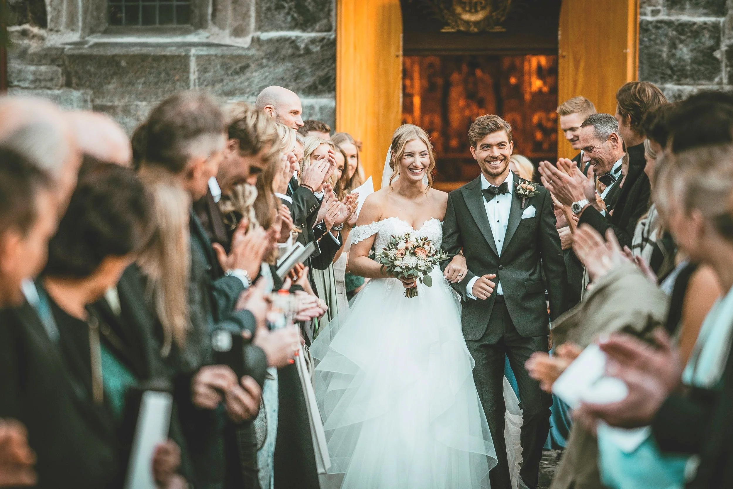 Bryllupsfoto/video: som går ut av en bygning, omgitt av klappende gjester i bryllupsklær.