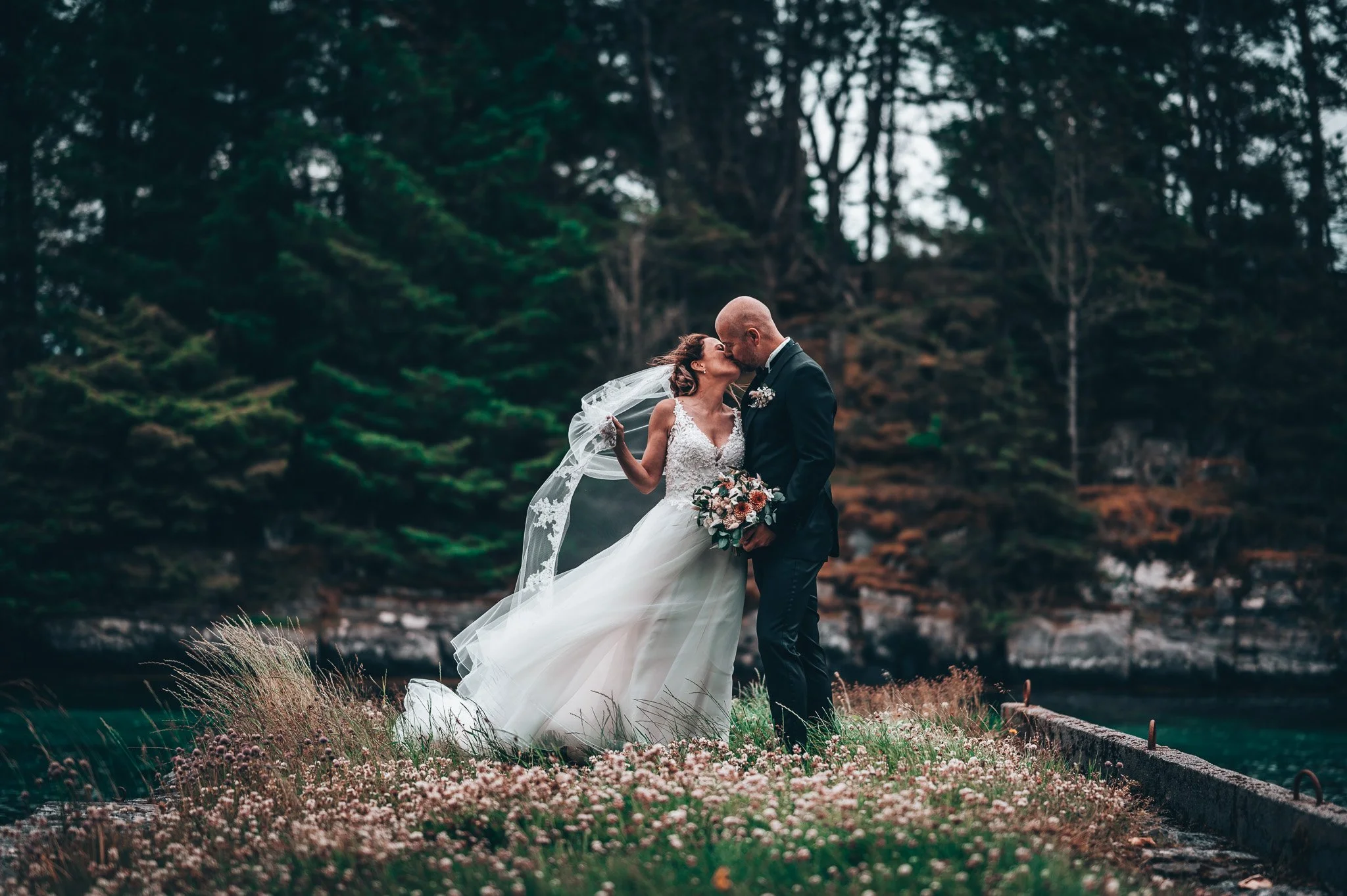 Brudepar kysser i skog med blomstereng, hun i hvit brudekjole med slør og blomsterbukett, han i mørk dress, omgitt av trær.
