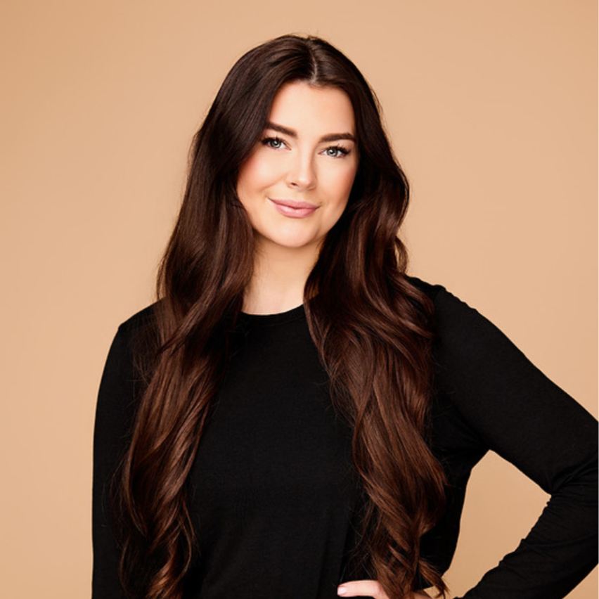 A woman with long brown hair in a black turtleneck smiles while holding an electric shaver in a room with neutral-colored walls and decor.