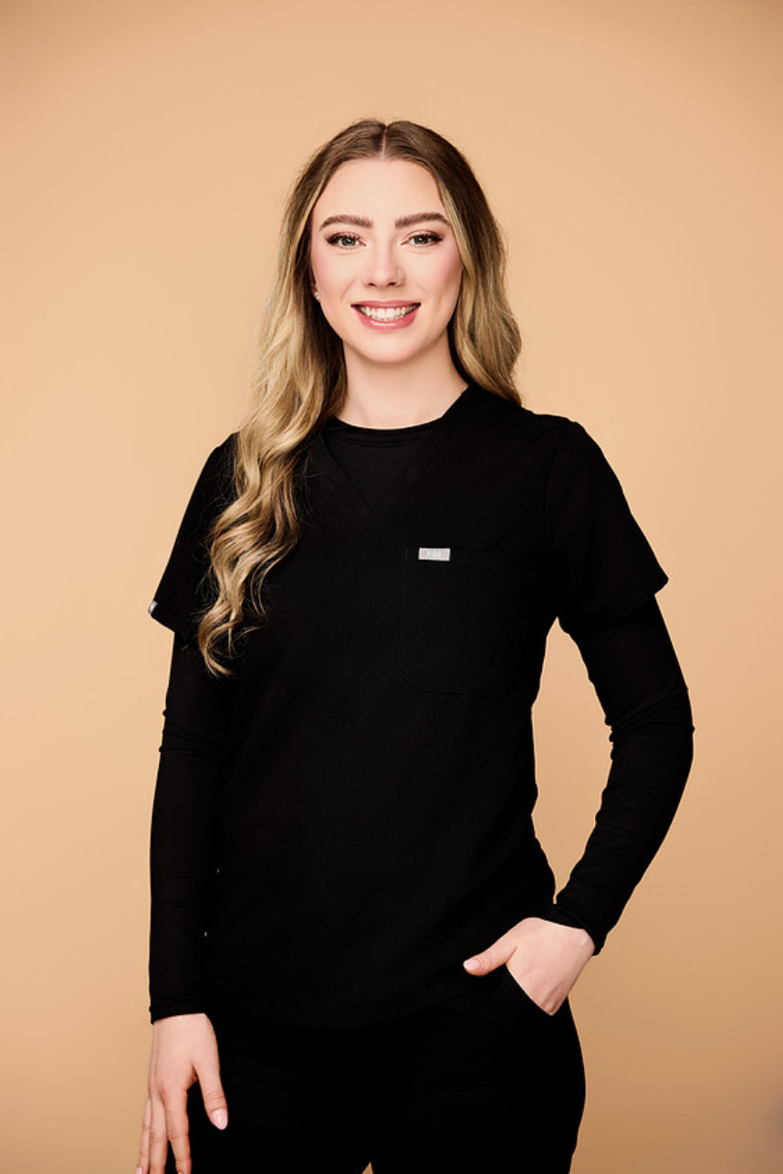 A smiling young woman with long blonde hair in loose waves, wearing a black long-sleeve shirt and black sweatpants with a gray drawstring, standing in a modern, well-lit reception area with a blurred logo in the background.