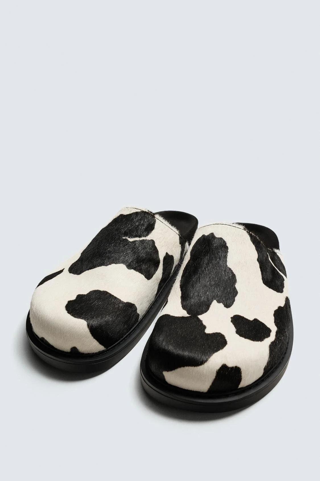 Paire de chaussons en imitation de peau de vache, noirs et blancs, sur un fond blanc.