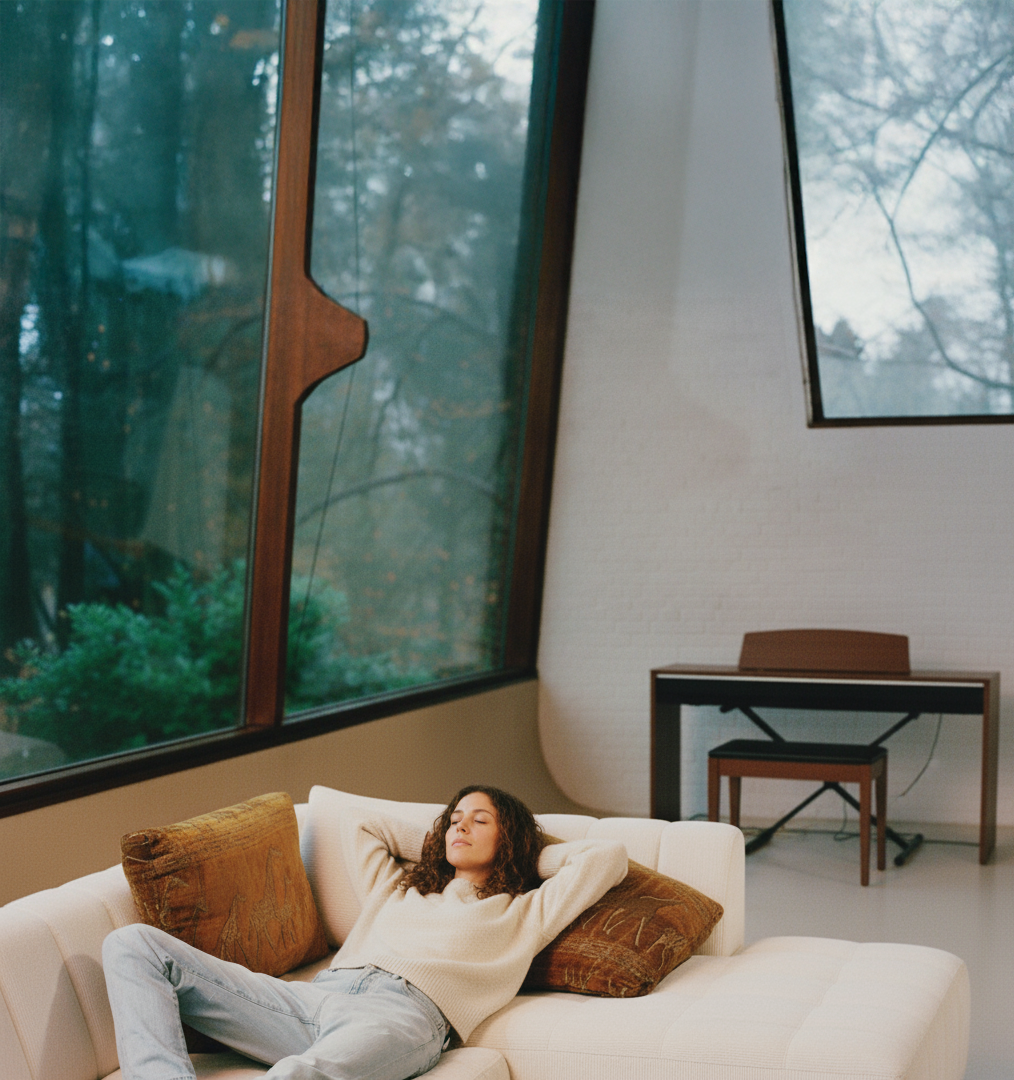 Visuel pub générée par IA d'une Jeune femme relaxant sur un canapé blanc à l'intérieur, avec des grandes fenêtres montrant une vue de la nature extérieure.