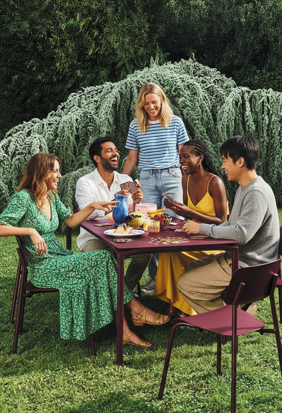 Un groupe de six personnes souriantes autour d'une table de pique-nique dans un jardin, partageant un moment convivial avec des gâteaux et des jeux.