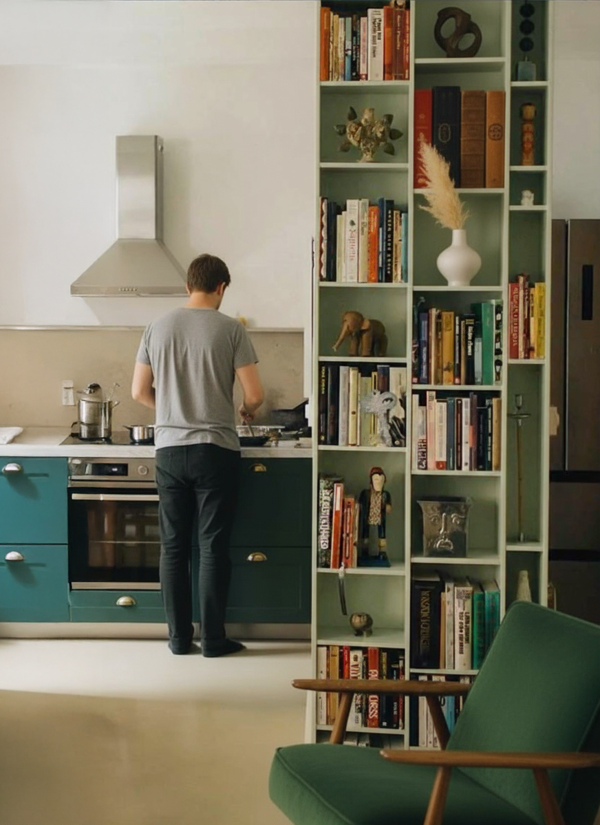Un homme prépare quelque chose dans la cuisine près d'une étagère remplie de livres et de décorations, vue partielle d'une pièce intérieure.