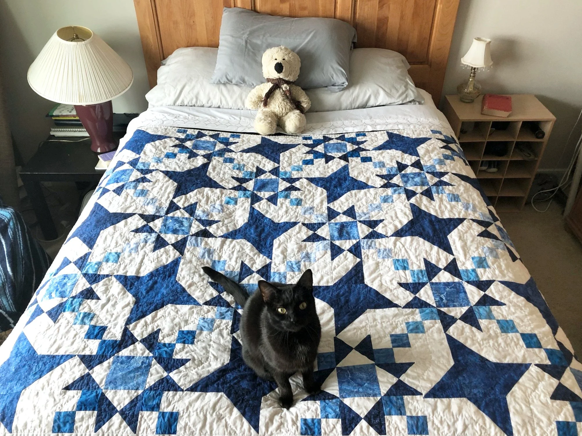 A full bed, nicely made, covered in a quilt of  blue and white stars in an alternating pattern. Also a black cat looking at the camera and a teddy bear resting on the pillows