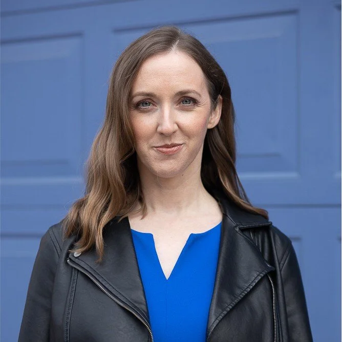 Headshot of Jennifer: white woman, early 40s, with light brown hair, a blue dress, and a black leather jacket; blue background