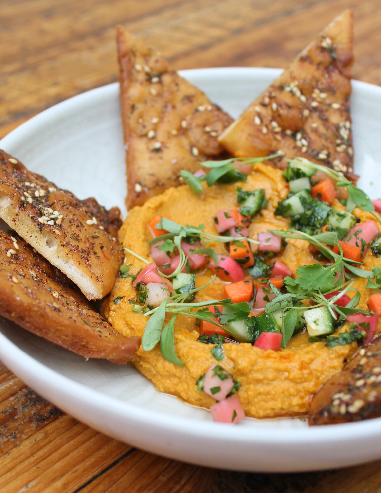 A bowl of vibrant orange Caramelized Carrot Hummus, topped with chopped cucumber, peppers, radishes, and fresh herbs. A serving of soft za'atar dusted pita chips is presented around the hummus.