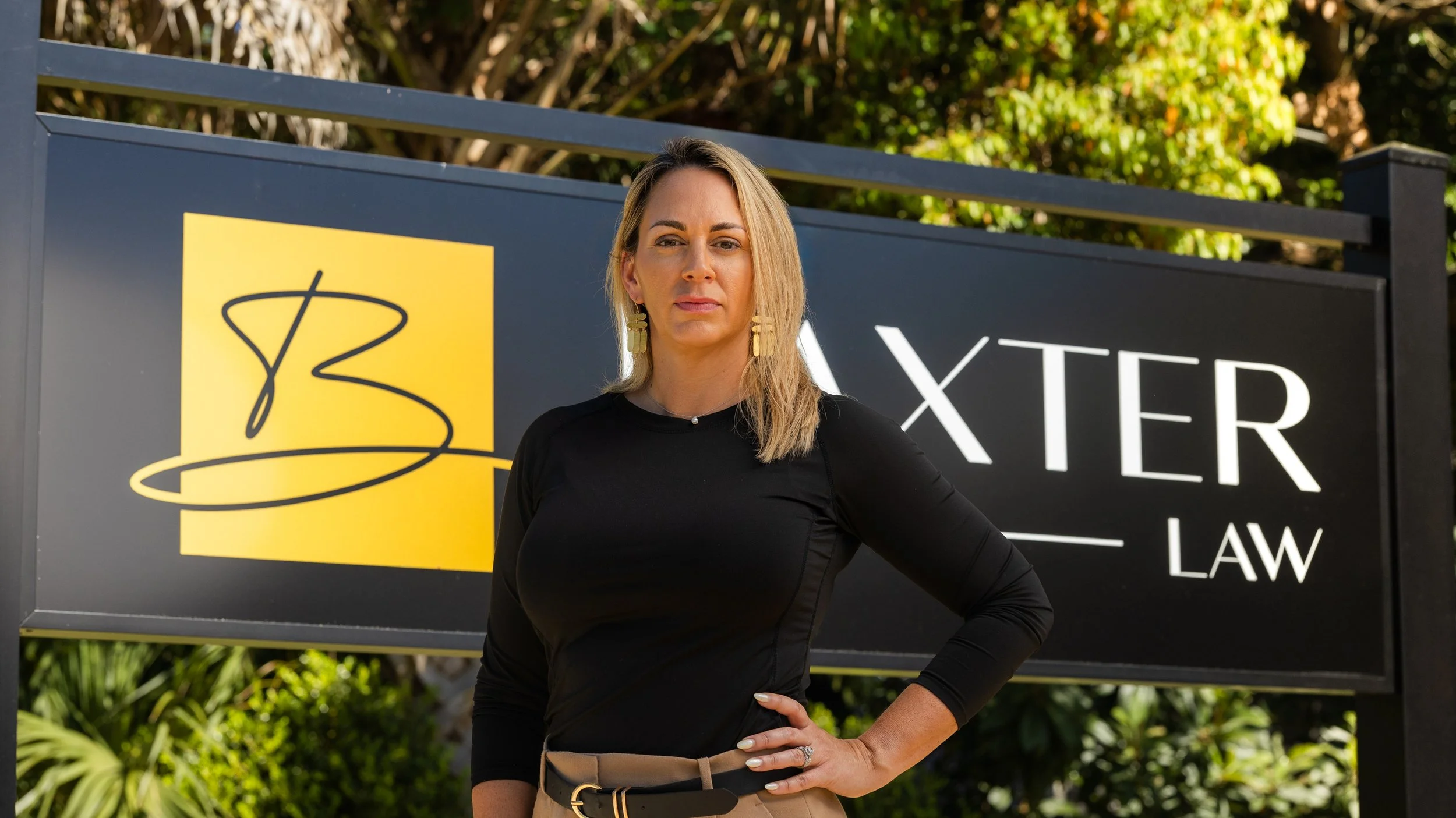 A woman with blonde hair, wearing a black long-sleeve top and beige pants, standing outdoors in front of a sign that reads 'BAXTER LAW' with a yellow logo, with green trees in the background.