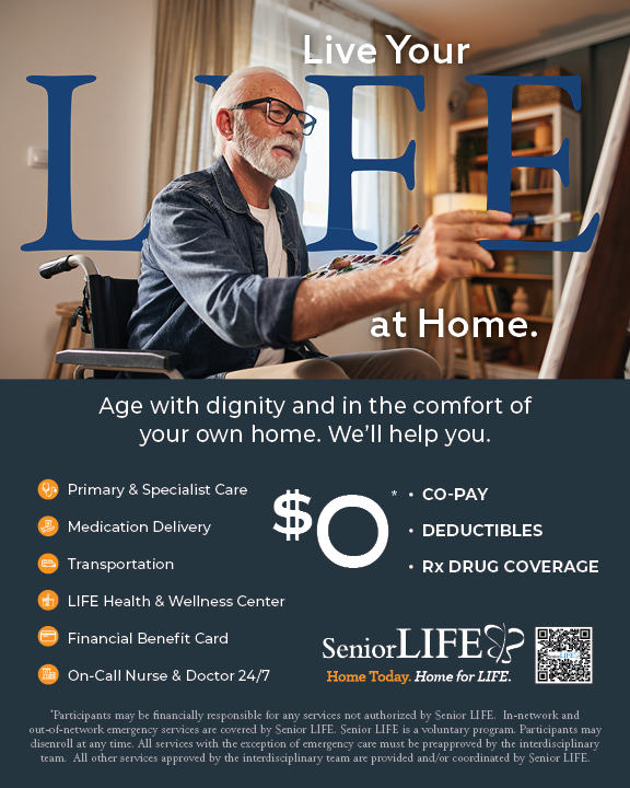 An elderly man with glasses and a beard painting on a canvas in a well-lit room with curtains and shelves containing books and decor.