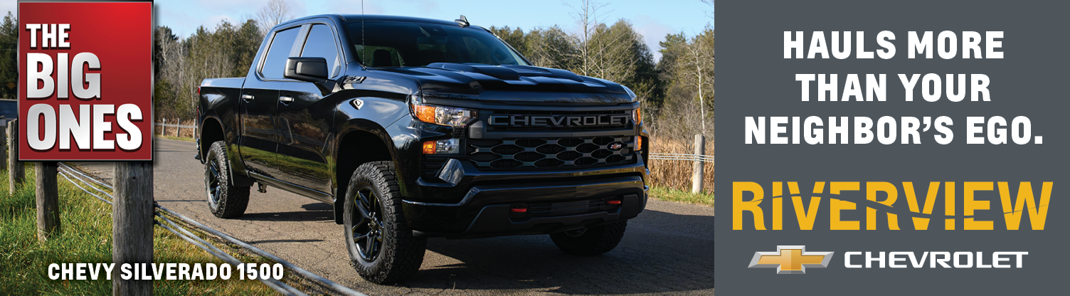 Black Chevrolet Silverado 1500 pickup truck parked on a roadside with trees in the background, advertisements for Riverview Chevrolet, and large text emphasizing the vehicle's size and features.