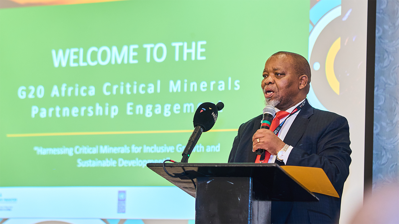 Man in suit speaking into a microphone at a conference with a green screen backdrop that reads 'Welcome to the G20 Africa Critical Minerals Partnership Engagement'.