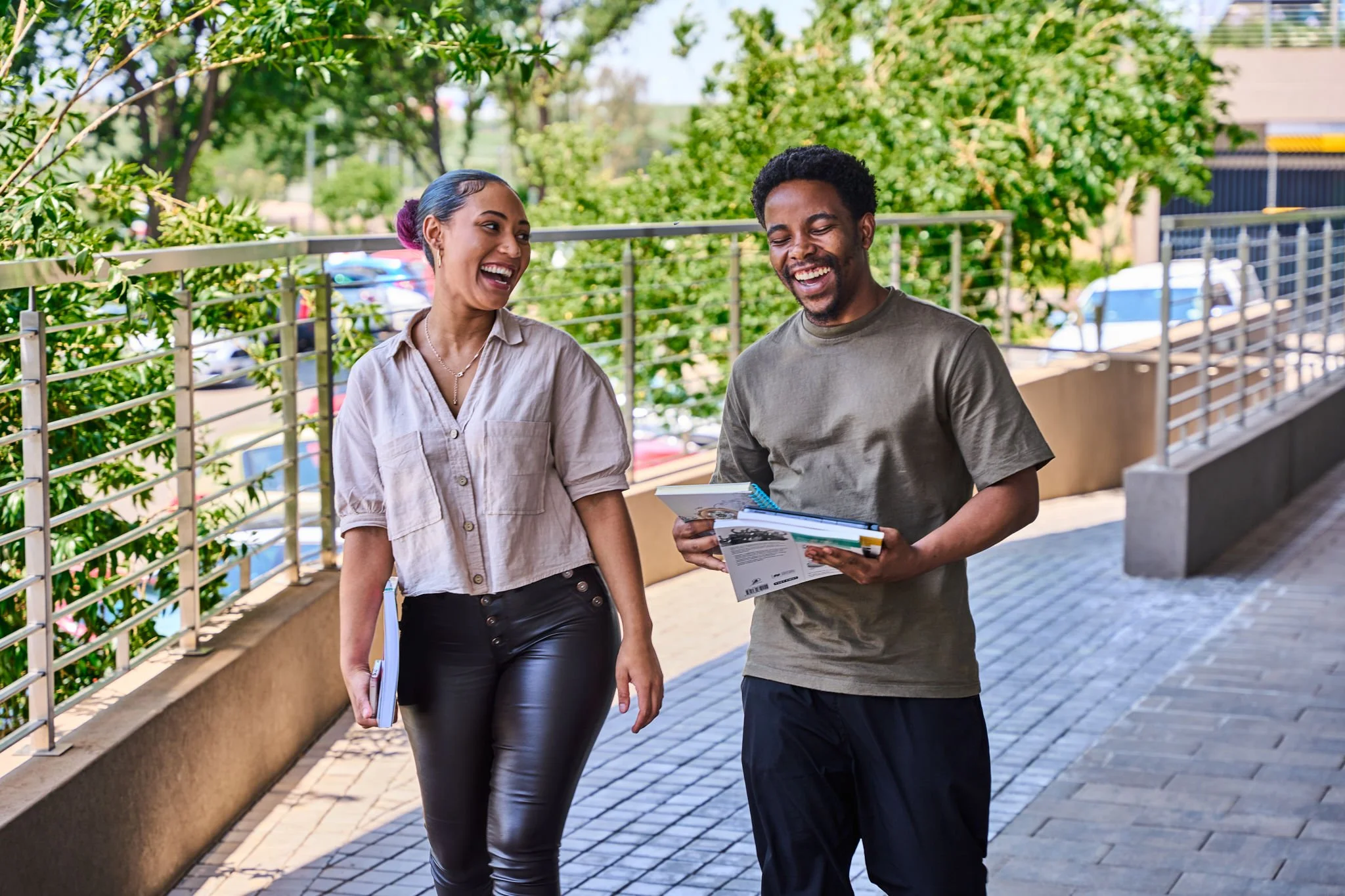A man and woman are walking and talking outdoors, both smiling. The man is holding pamphlets or brochures, and the woman is carrying a notebook or tablet. They are on a paved walkway with trees and a metal railing in the background.