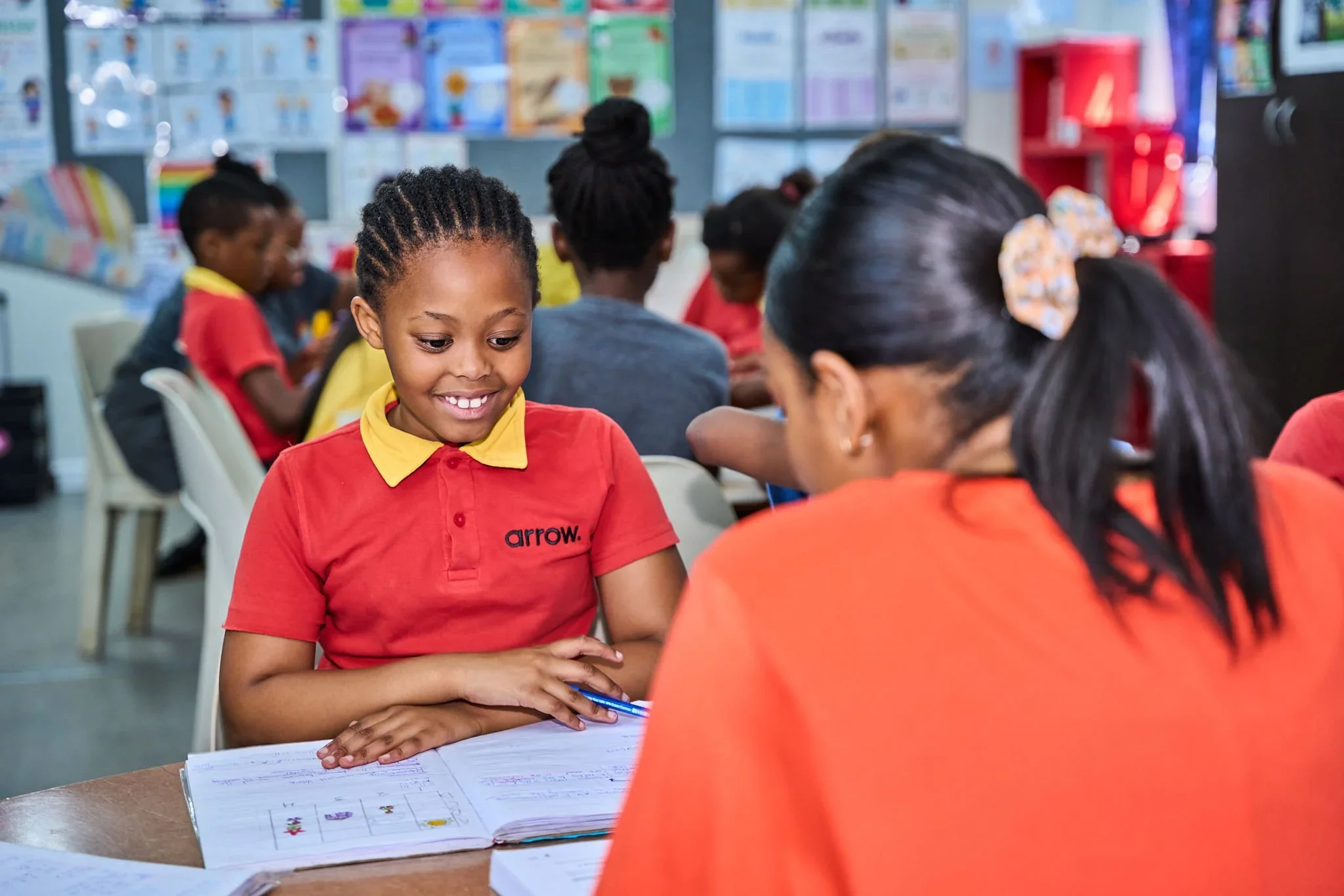 A young girl in a red and yellow school uniform smiles at a teacher while looking at her workbook in a classroom. Other students are seated at tables in the background.