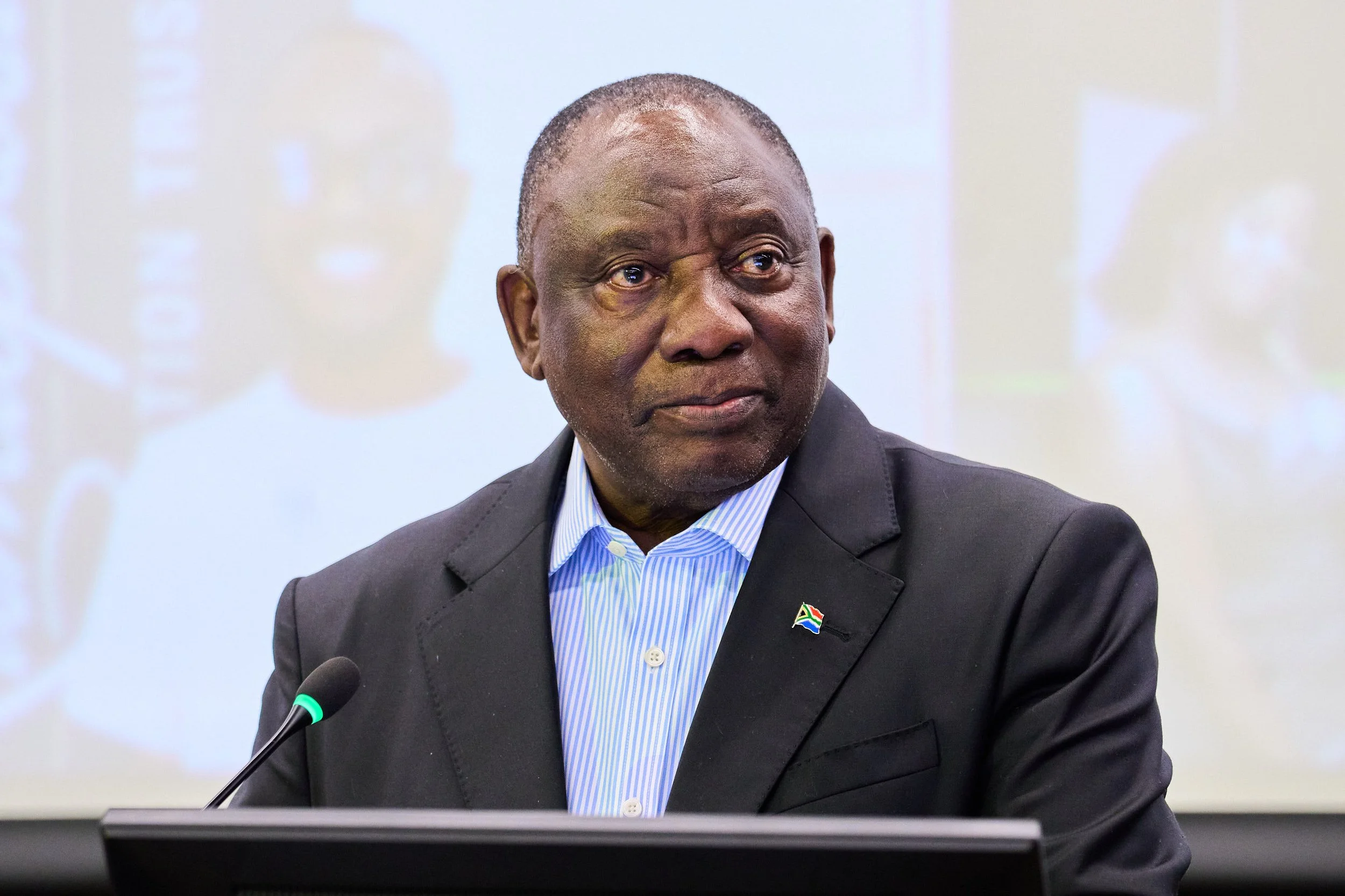 An older man with dark skin, short gray hair, wearing a dark suit, light blue striped shirt, seated at a table with a microphone, with a blurred background showing a screen or projection.