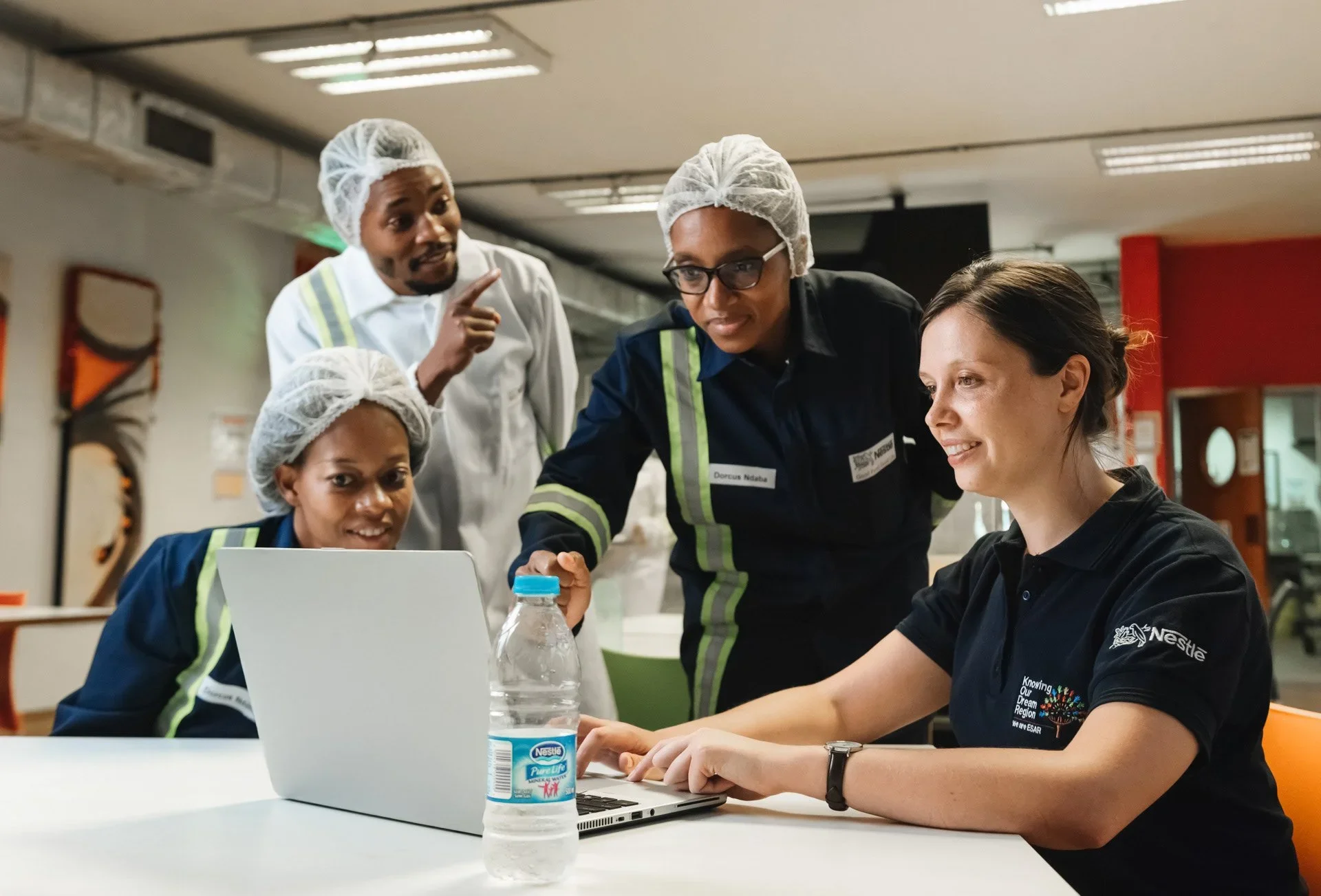 Four people, two women and two men, in a workplace, wearing hairnets and work uniforms, gathered around a laptop, engaged in a discussion.