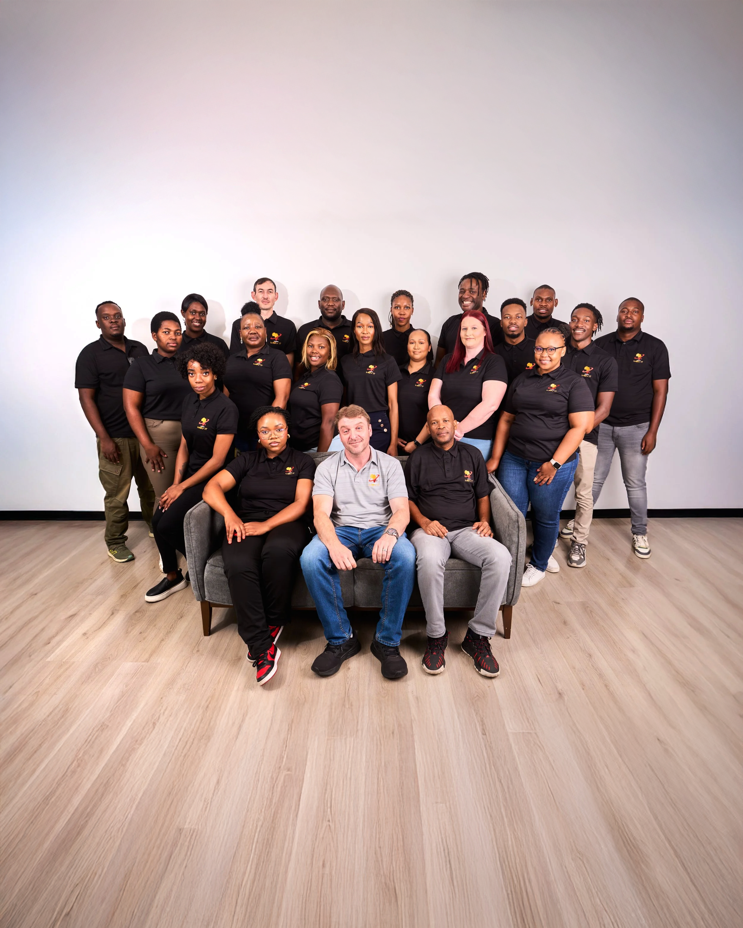 Group of 16 diverse people, some seated and some standing, posing in a studio with a plain white background, wearing black polo shirts with a colorful logo.