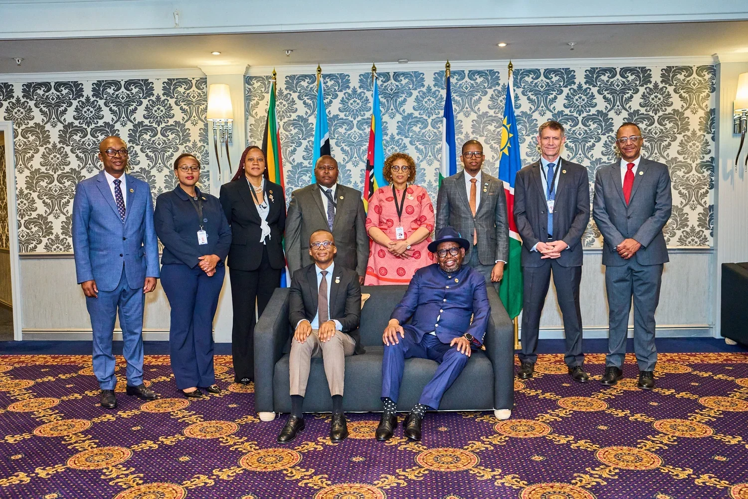 Group of nine people, six men and three women, in formal business attire, standing and sitting in front of a backdrop of national flags and patterned wallpaper.