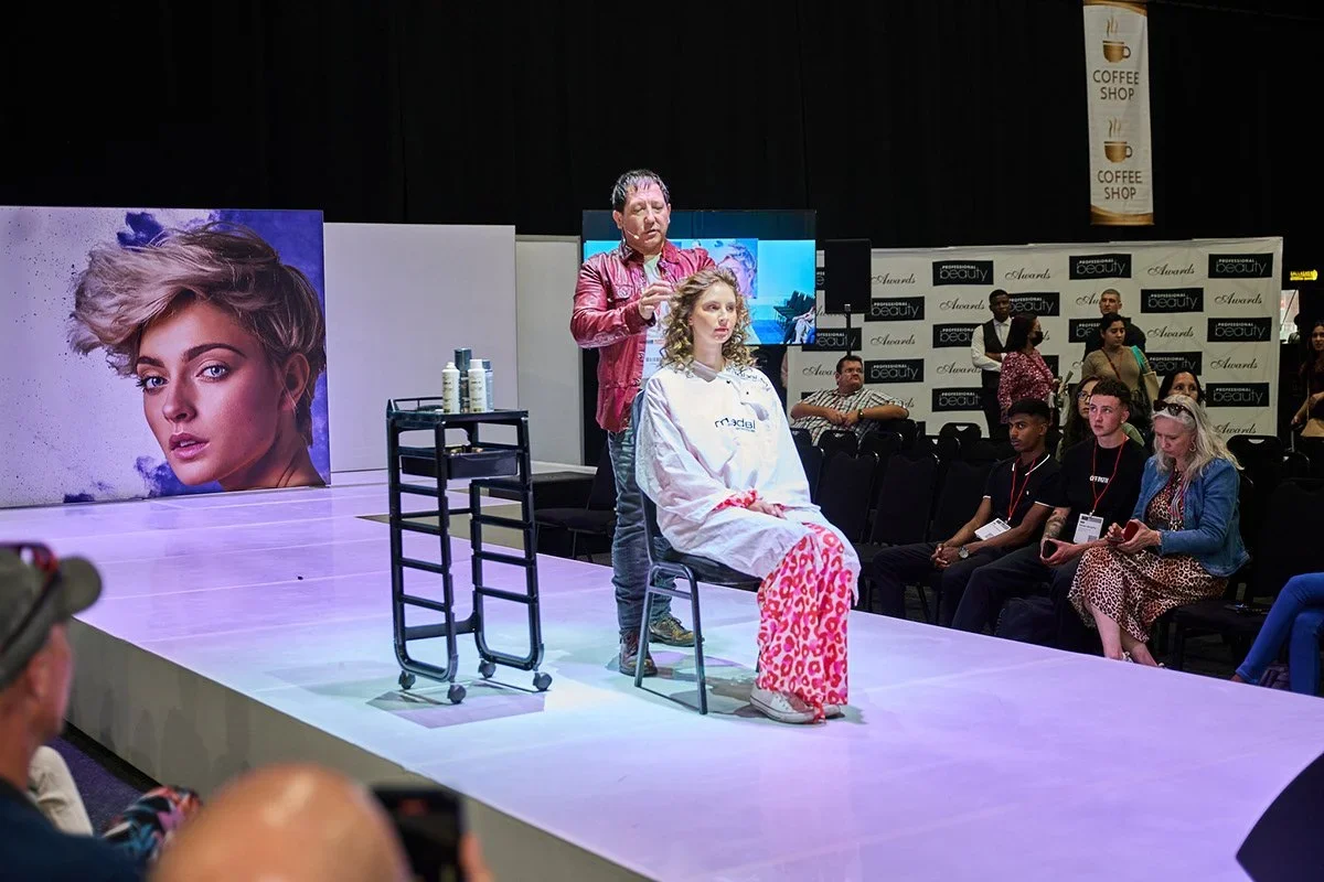 Hairdresser cutting a woman's curly hair on a stage in a public event, with beauty product displays and audience seated nearby.