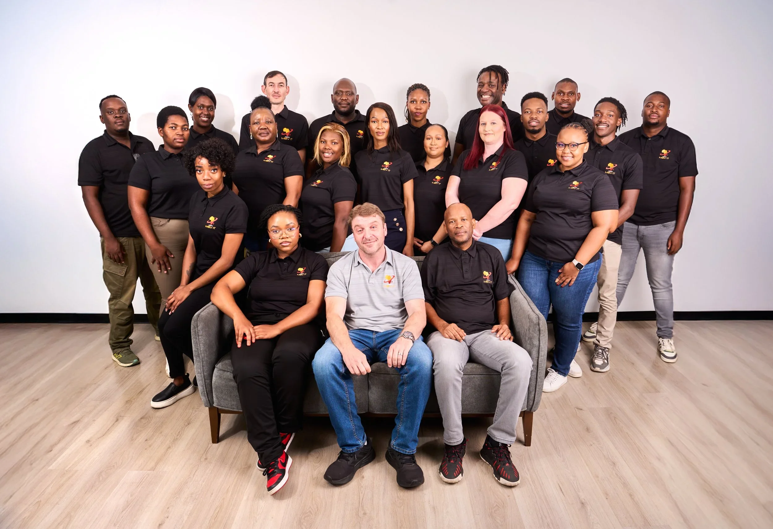 A diverse group of 18 people posing for a photo indoors against a plain white wall, with some seated on a gray sofa and others standing behind. Everyone is wearing casual black shirts, some with a colorful logo on the chest.